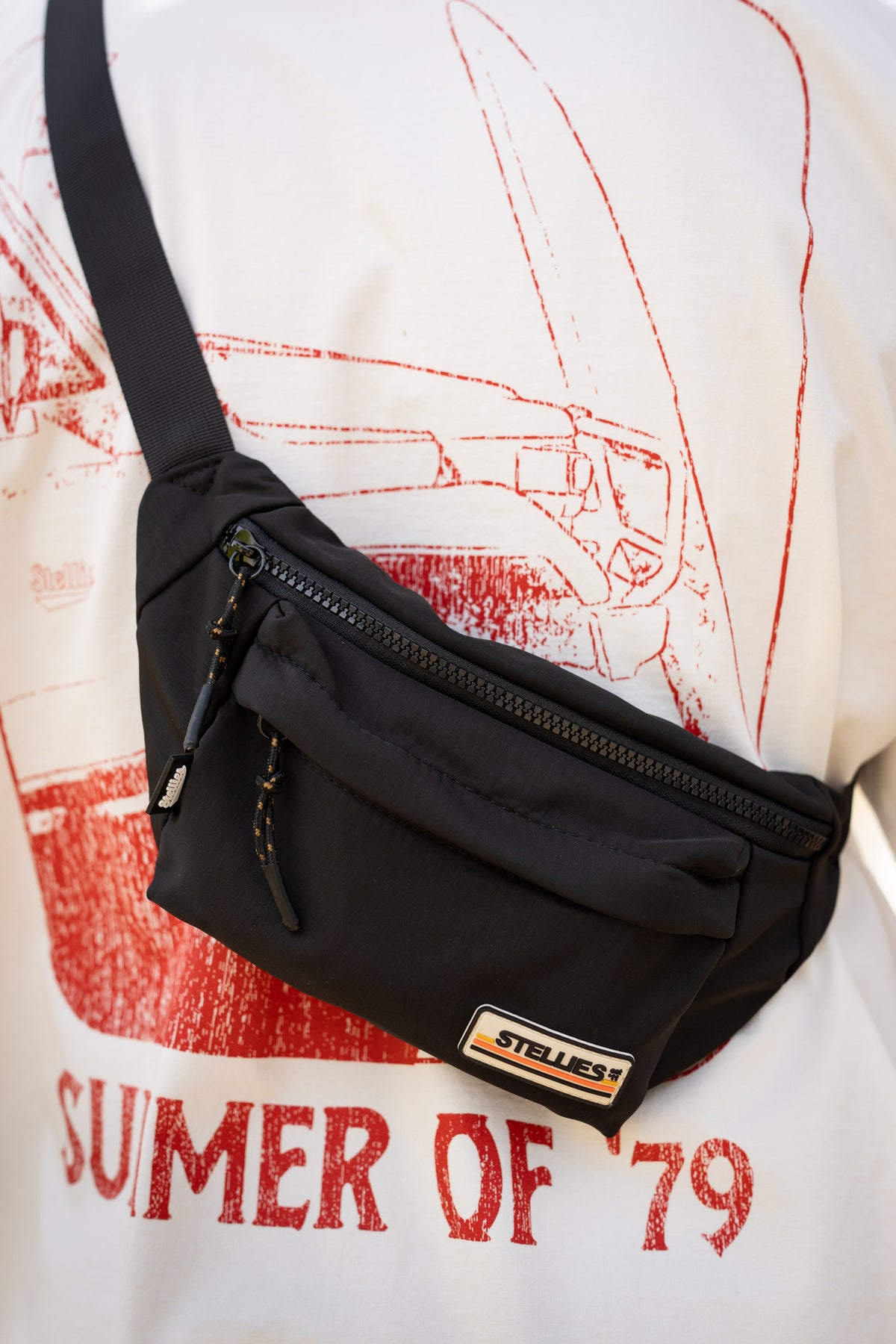 A black nylon crossbody fanny pack with a zippered front pocket and adjustable shoulder strap, worn across a white graphic t-shirt with red print. The brand name 'STELLIES' is visible on a small rectangular patch on the front of the bag.