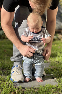 Person helping a toddler stand on a stone path in a grassy area.